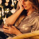 une femme avec sa tête dans sa main qui a l'air bouleversée par un arbre de Noël tenant un verre de vin dans son autre main